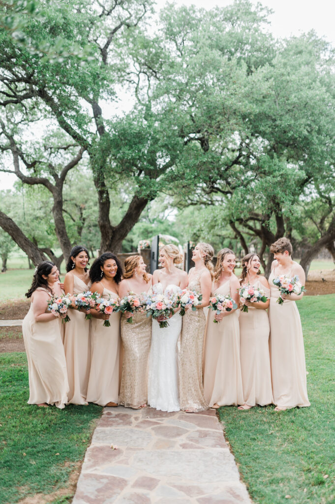Bride with bridesmaids in neutral dresses holding colorful floral bouquets beneath oak trees.