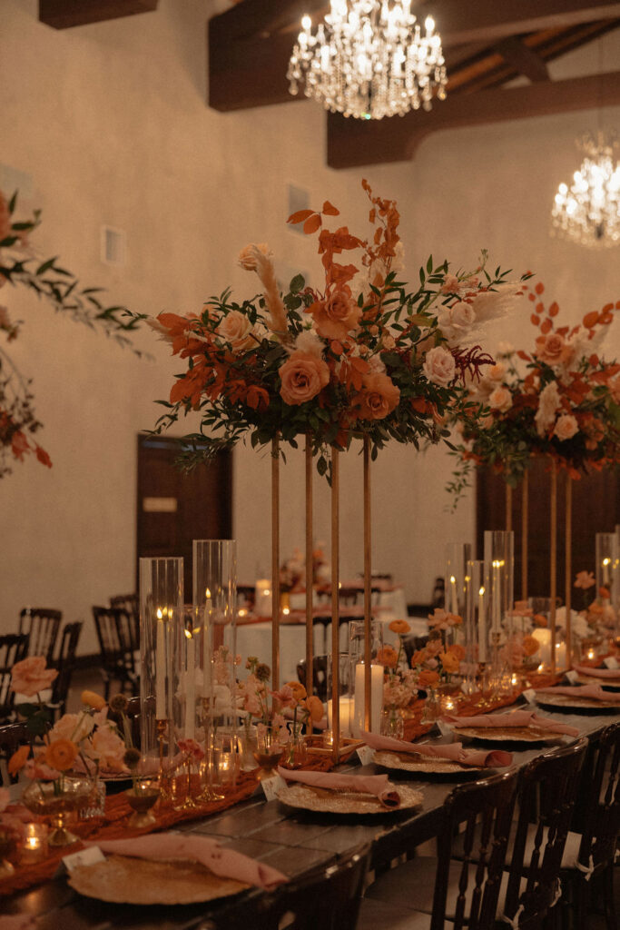 Tall orange and blush floral centerpieces decorating a long wedding reception table.
