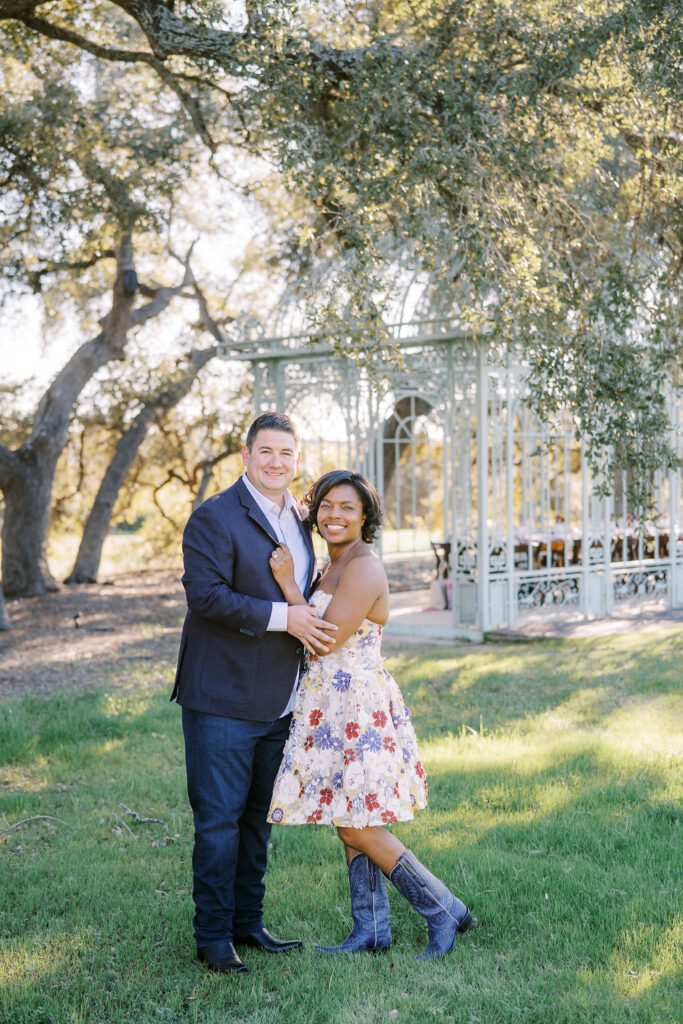 A couple poses on green grass with a gazebo behind them, reflecting Engagement Party Etiquette in 2026