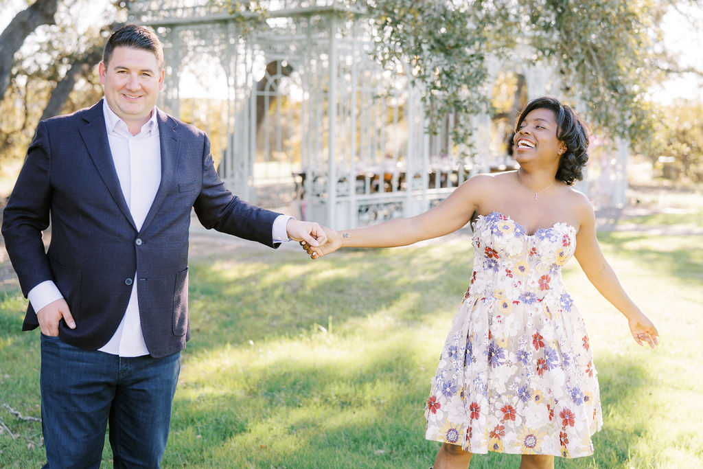 A couple holds hands and smiles in front of an elegant outdoor setting, depicting Engagement Party Etiquette in 2026