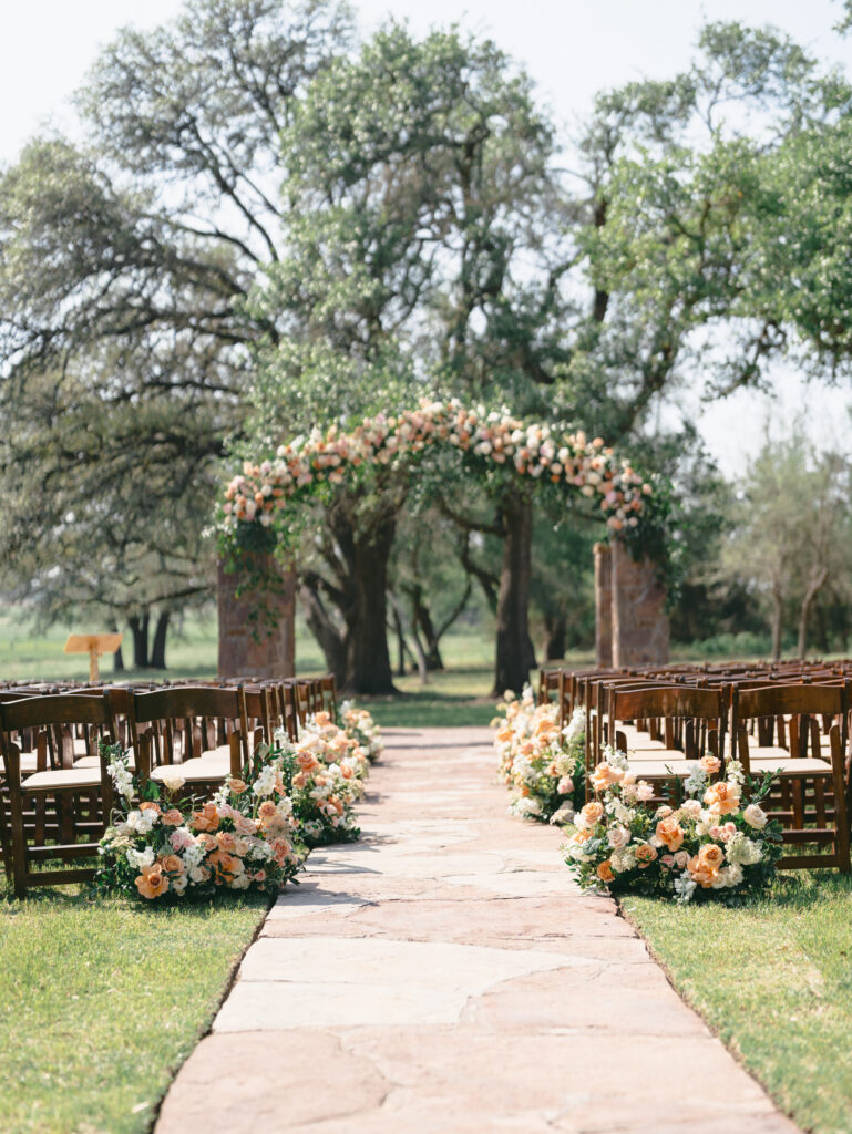Outdoor wedding ceremony aisle lined with peach flowers and a floral arch.
