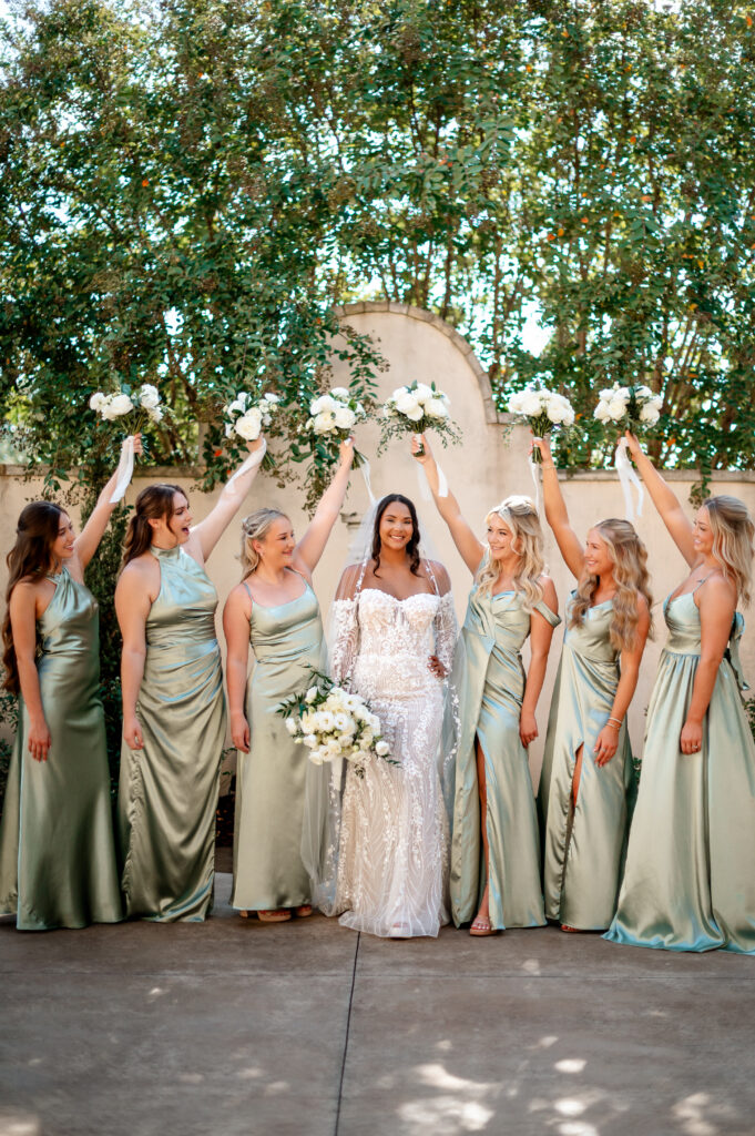 Bride with bridesmaids in sage green dresses raising white floral bouquets.