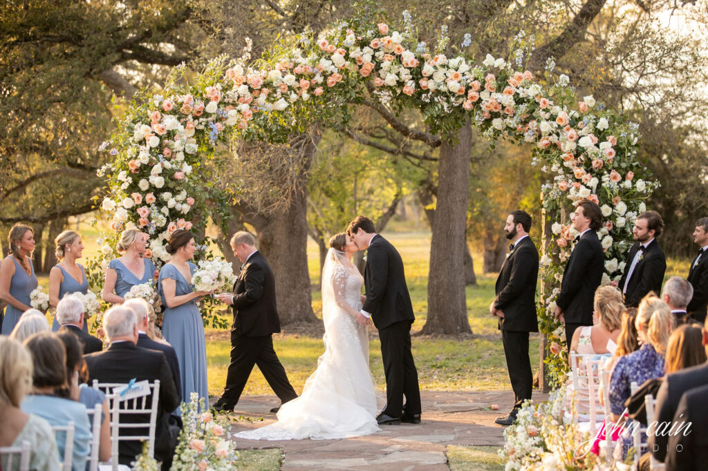 bride and groom kissing under coral and white floral arch at Ma Maison wedding venue