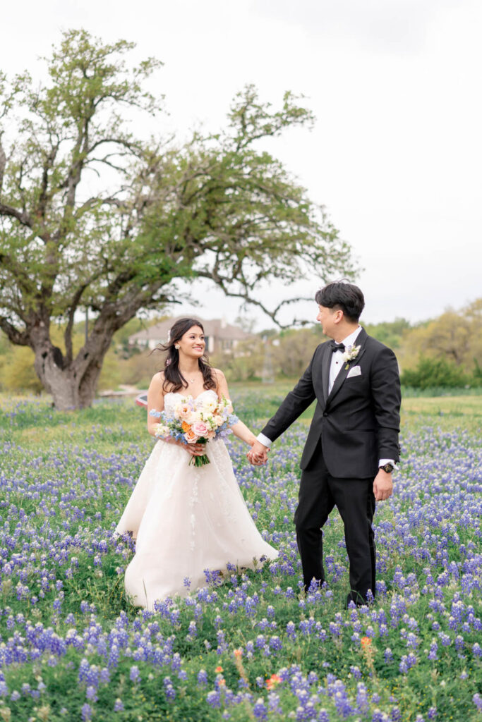 A newlywed couple holds hands in a field of Texas bluebonnets, captured beautifully by an Austin videographer at Ma Maison