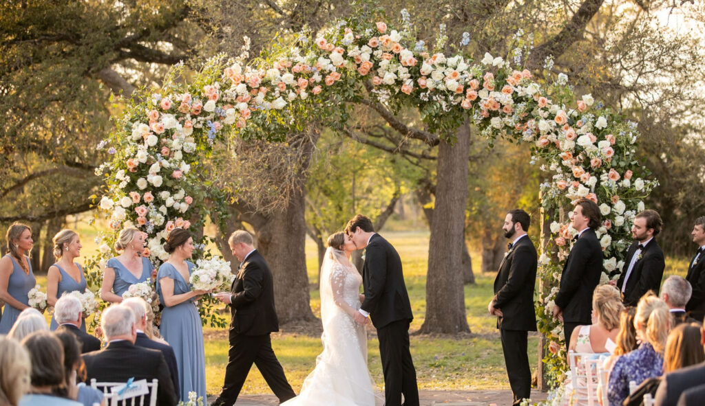 Couple’s first kiss beneath a floral arch at an Austin wedding venue with European elegance