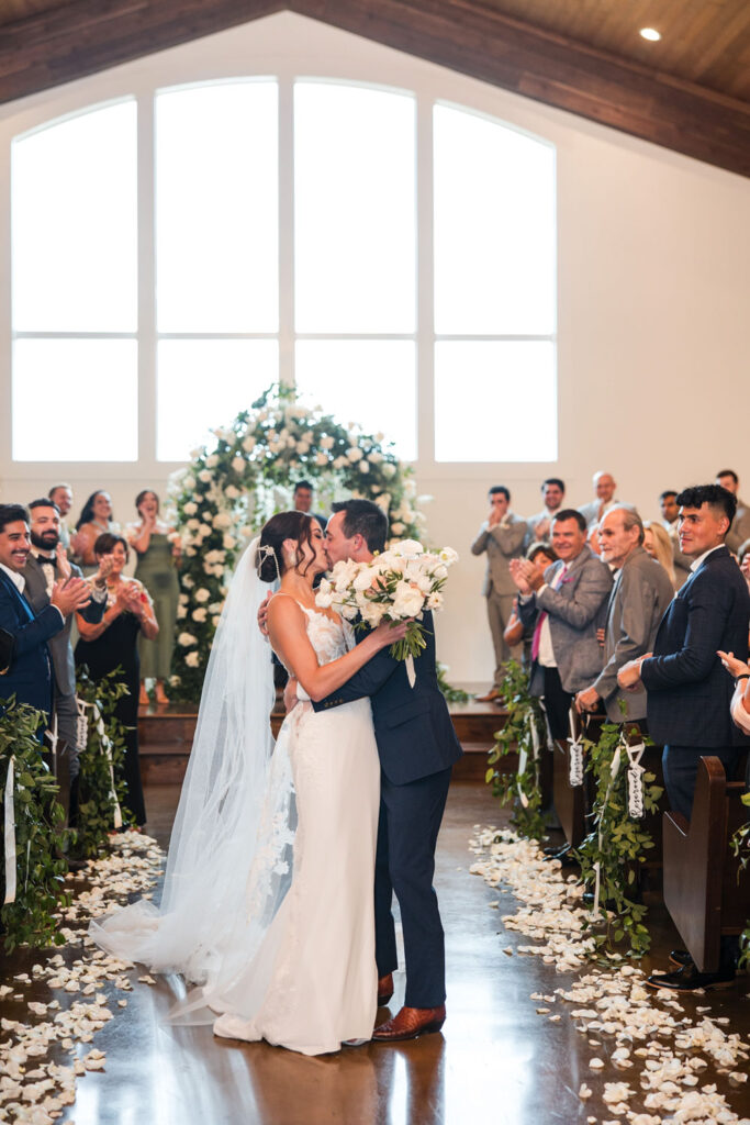 A newlywed couple shares their first kiss as husband and wife in a breathtaking indoor ceremony at Ma Maison, the best Austin wedding venue for a rainy day