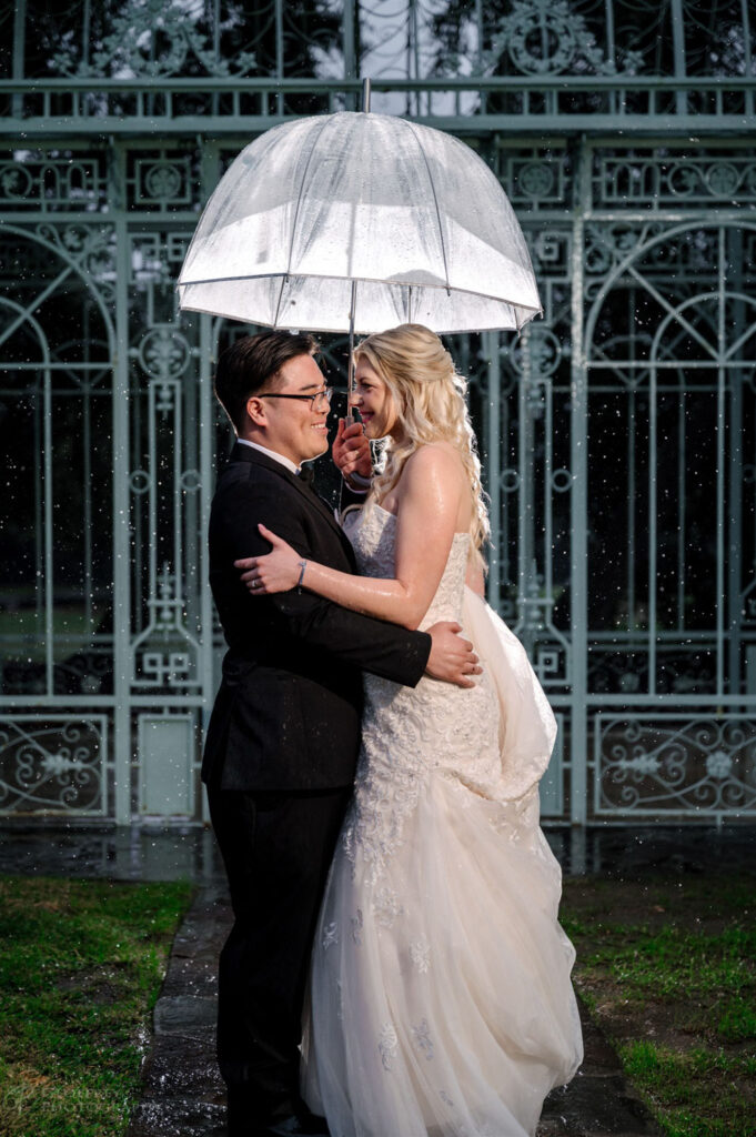 A bride and groom share a cozy embrace under an umbrella at Ma Maison’s scenic outdoor structure