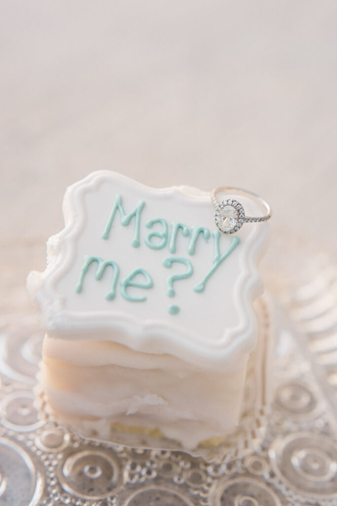 Mini engagement cake with “Marry me?” written in icing and topped with a diamond ring.