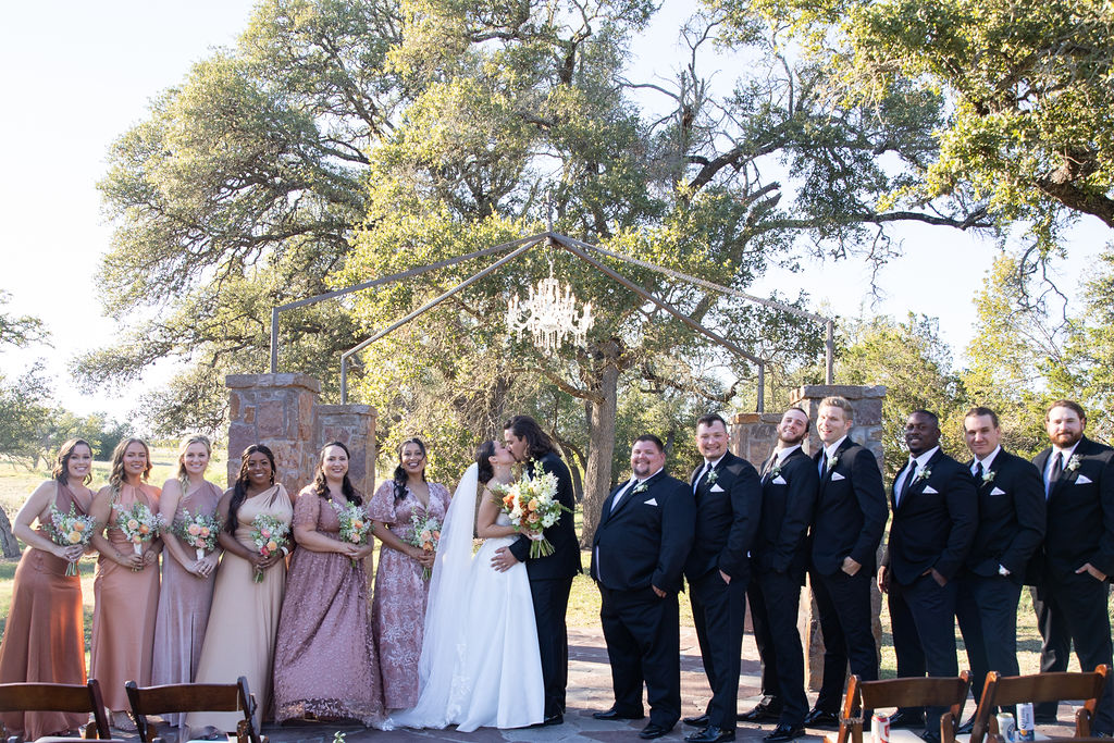 Bride and groom kissing with wedding party in black-tie wedding attire outdoors