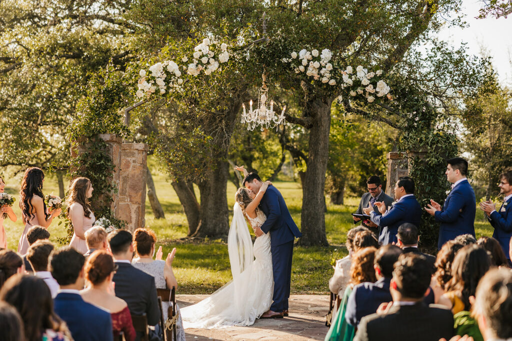 Newlyweds sharing their first kiss under a floral arch at an outdoor Dallas wedding venue near Austin