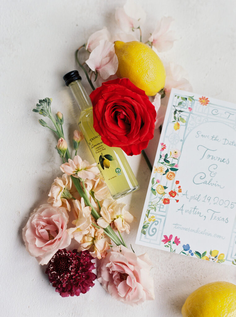 Colorful wedding stationery flat lay with flowers, lemons, and favor bottle.