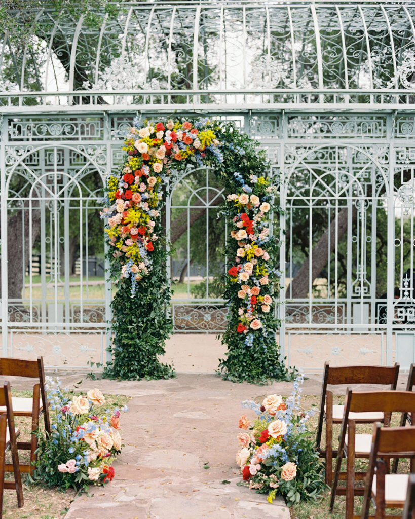Floral wedding ceremony arch in front of an ornate glass greenhouse.