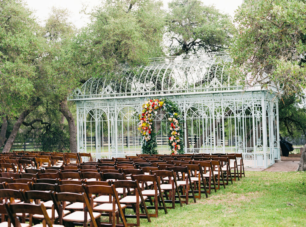 Outdoor wedding ceremony setup with wooden chairs and floral greenhouse backdrop.