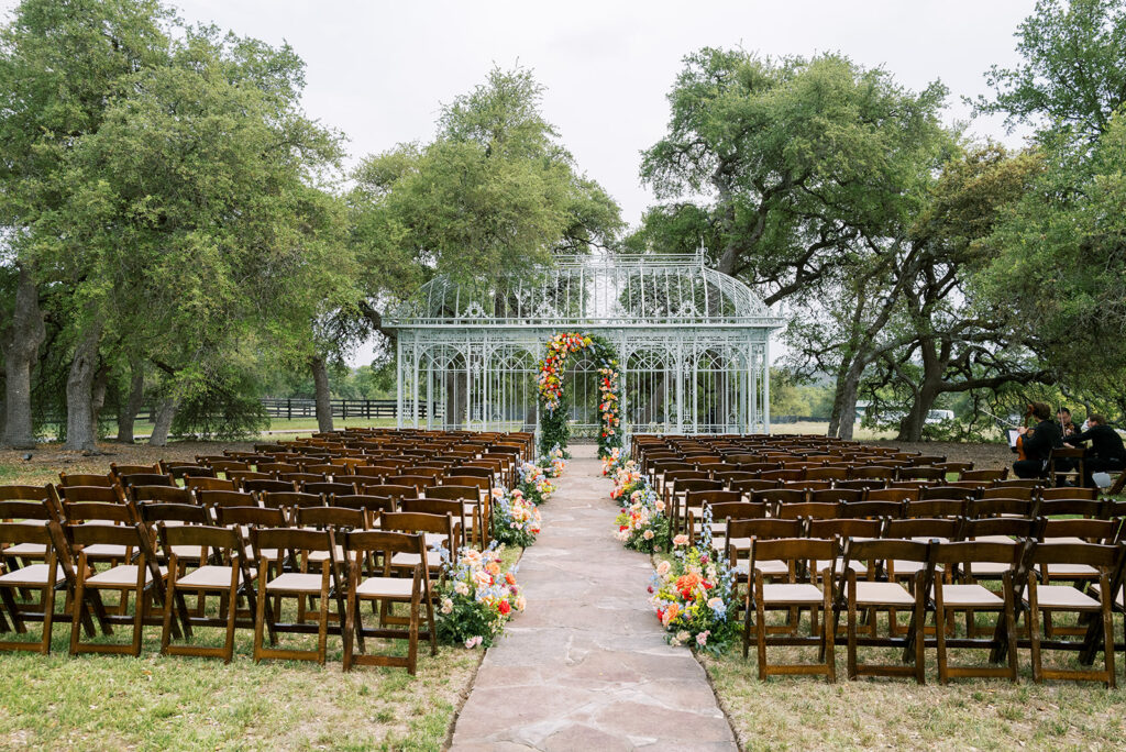 Outdoor wedding ceremony with wooden chairs, colorful aisle flowers, and greenhouse backdrop.
