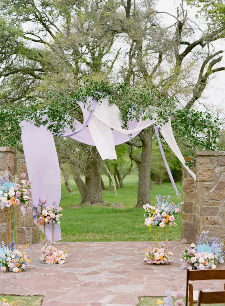 Outdoor ceremony arch draped in lavender fabric with pastel floral arrangements at Ma Maison