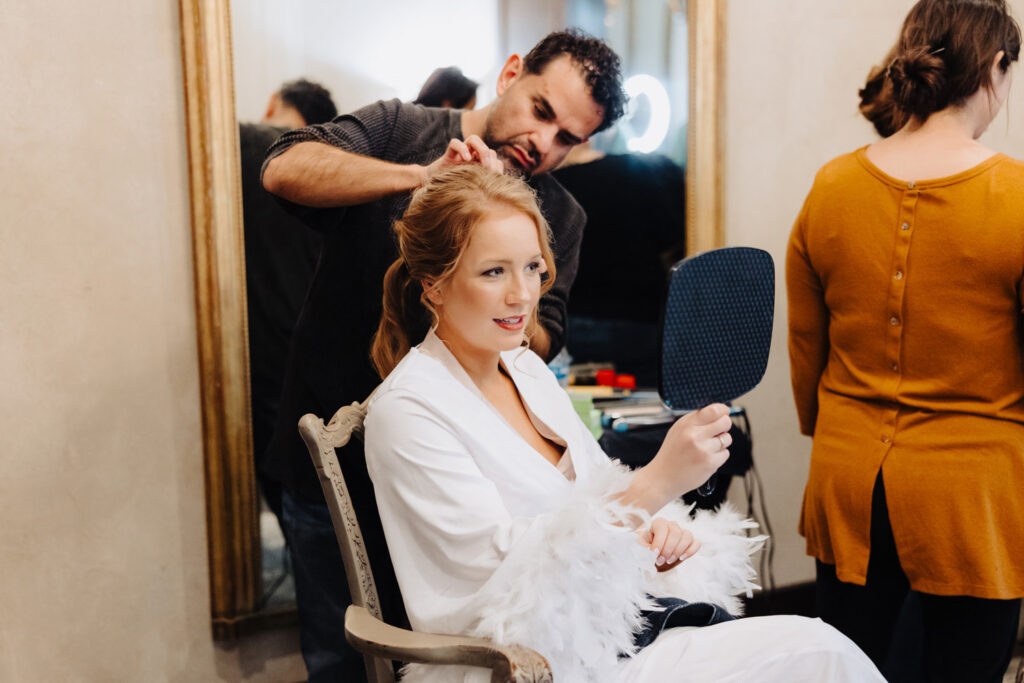 Bride sitting in a vintage chair admiring her look in the mirror while getting her hair styled by a professional hair and makeup artist.