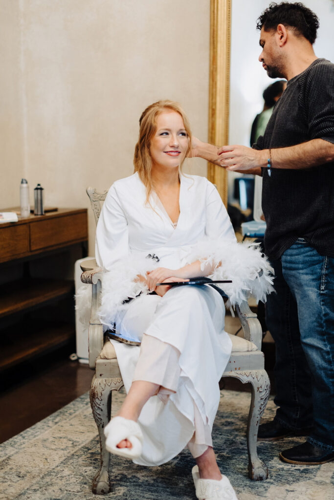 Bride in a feather-trimmed robe with ‘Mrs’ slippers, enjoying her glam session with a talented wedding hair and makeup artist in Austin.