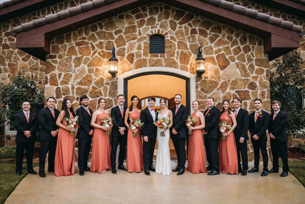 Bridal party in burnt orange dresses and black suits in front of stone chapel at Austin Italian wedding.