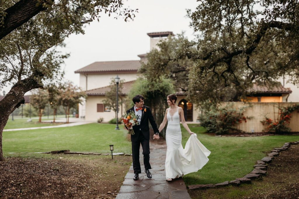 A newlywed couple walks hand in hand through the lush gardens of Ma Maison, a premier Tuscan Destination Wedding Venue, featuring elegant European architecture