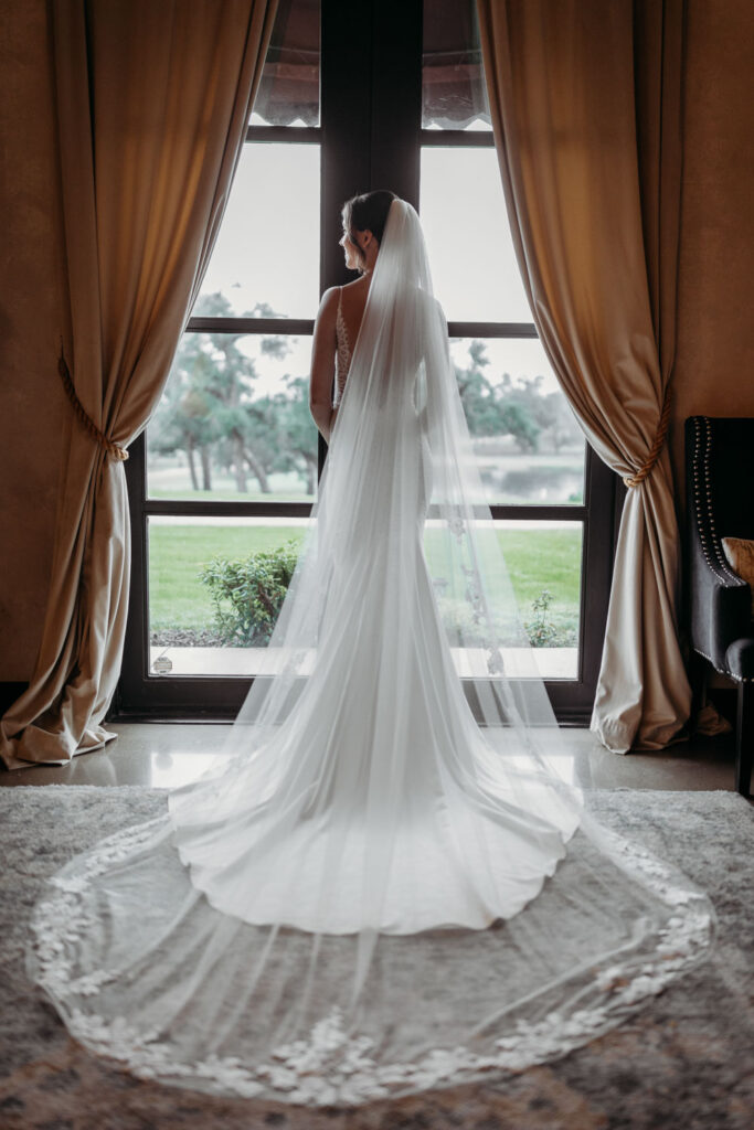 A bride stands gracefully by a sunlit window at Ma Maison, a Tuscan Destination Wedding Venue, her flowing veil cascading to the floor in timeless elegance