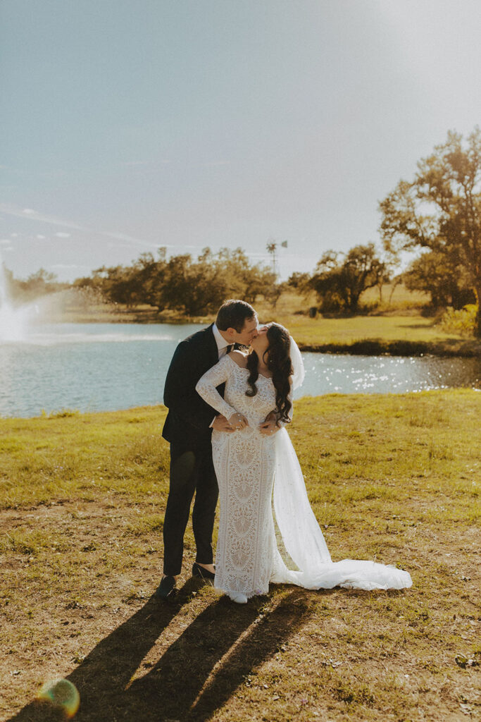 A bride and groom share a romantic kiss by the lake at Ma Maison, a Tuscan Destination Wedding Venue, with golden sunlight reflecting off the serene water.