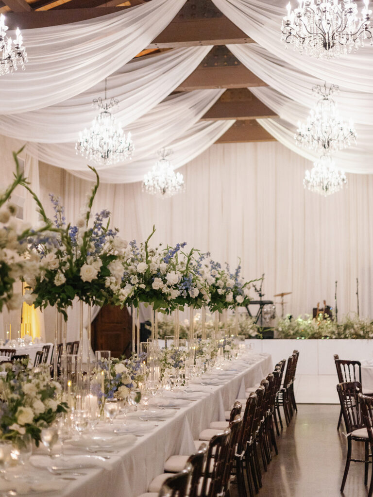 Long wedding reception table with white and blue florals beneath draped fabric and chandeliers.
