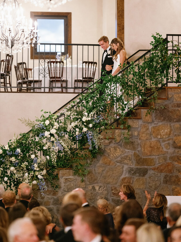 Bride and groom standing on a stone staircase decorated with greenery and white flowers.