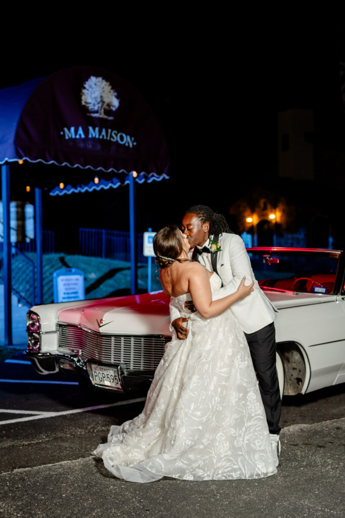 The newlyweds share a kiss beside a classic car under the Ma Maison awning at night