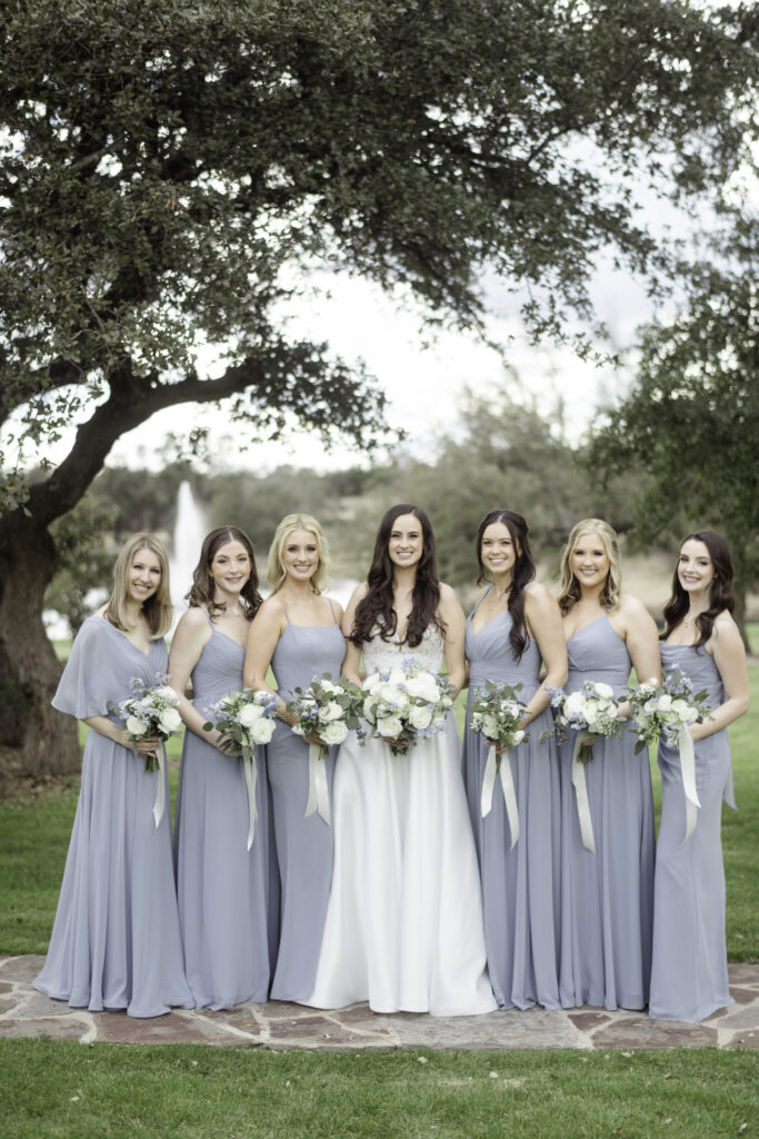 Bride with bridesmaids in dusty blue dresses holding floral bouquets.