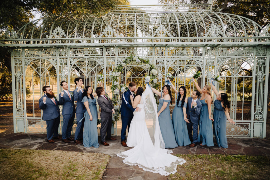 Bride and groom share a joyful kiss surrounded by their wedding party under a romantic floral arch at a Tuscan-inspired venue