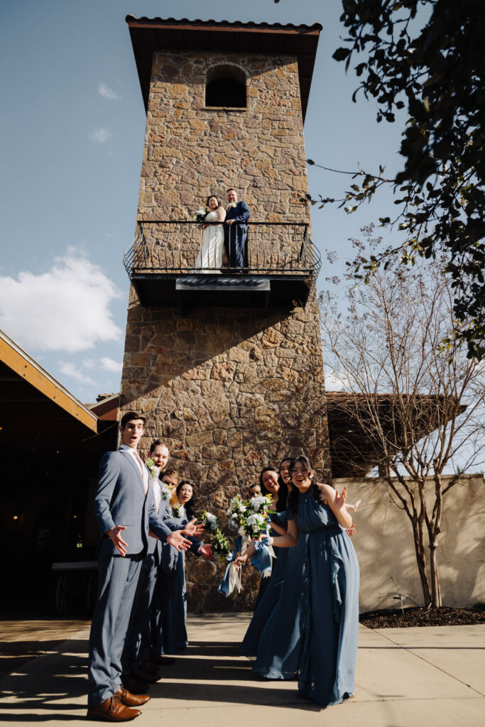 Bridal party celebrates below as the couple waves from a rustic stone tower at their Tuscan wedding venue