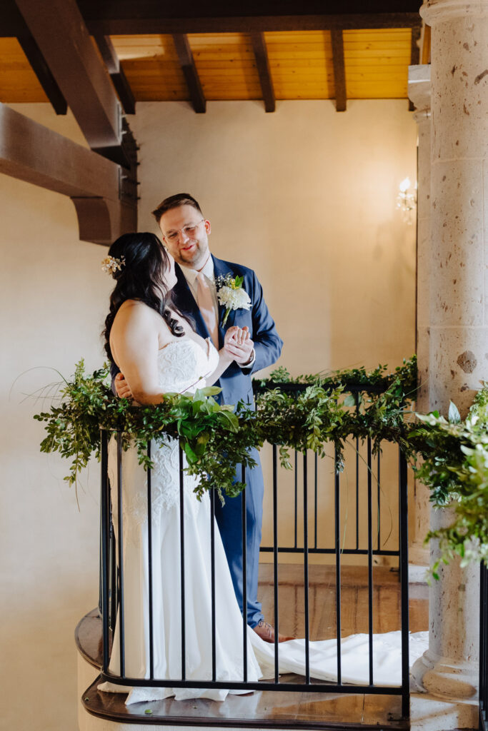 Bride and groom holding hands on a curved indoor staircase wrapped in greenery