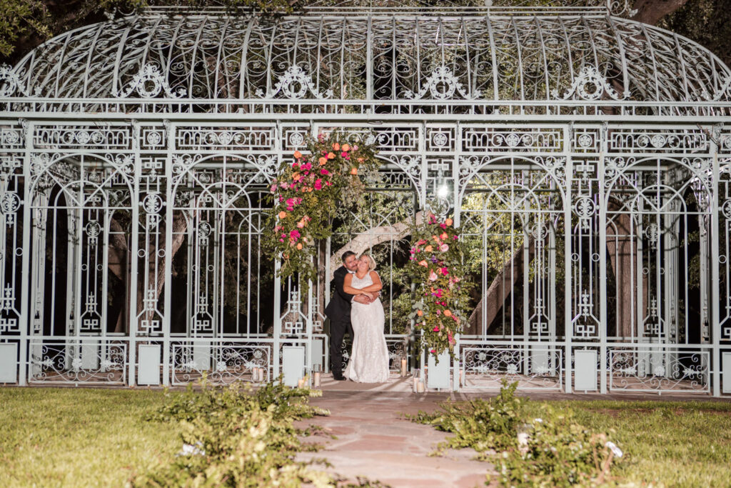 Newlyweds smiling in front of a floral arch at their outdoor wedding venue