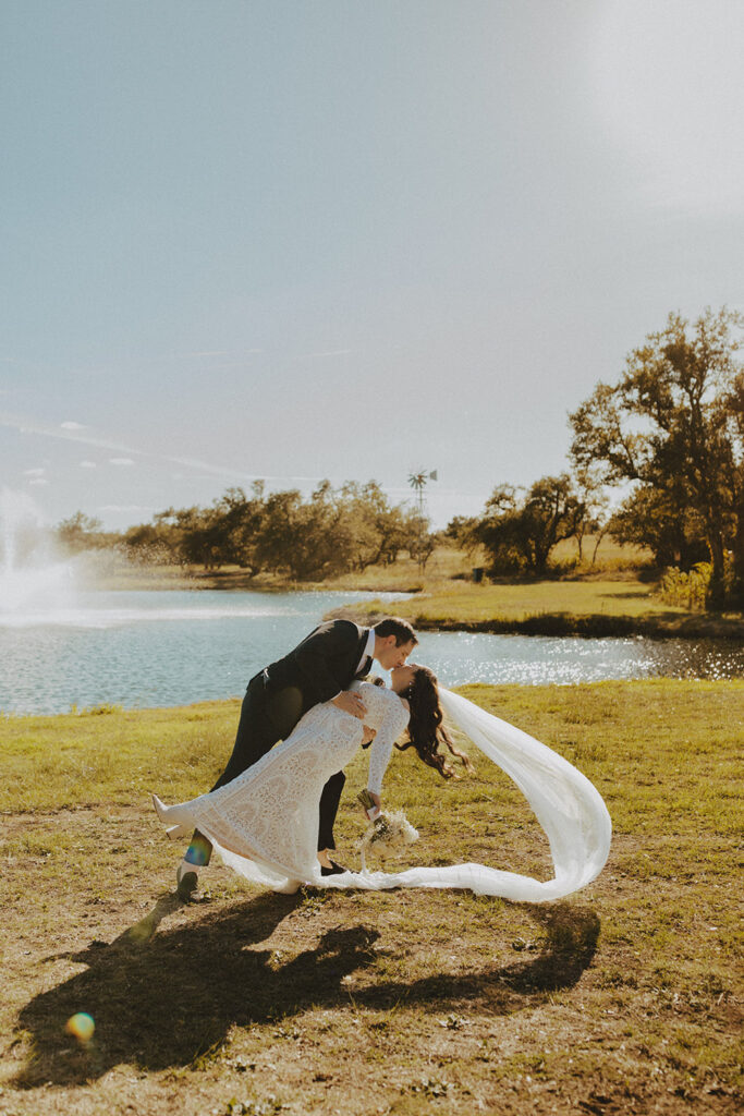 Groom dip kissing the bride beside Ma Maison’s lake under bright sun