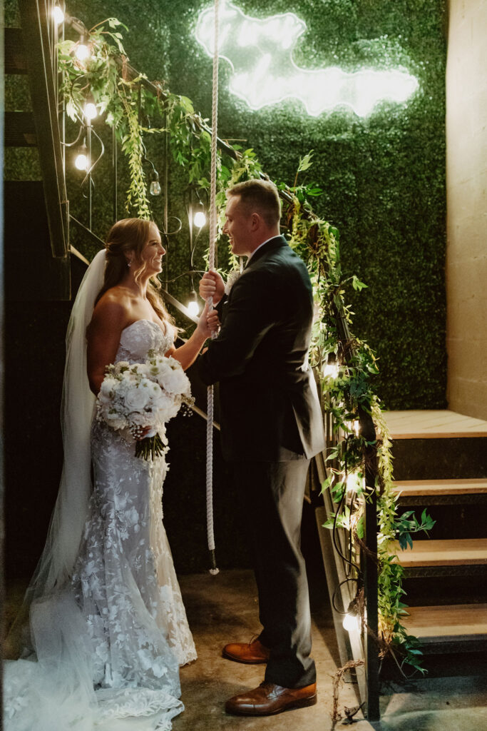 A romantic indoor wedding ceremony at Ma Maison in Austin, TX, with the bride and groom sharing a heartfelt moment under soft, glowing string lights and lush greenery