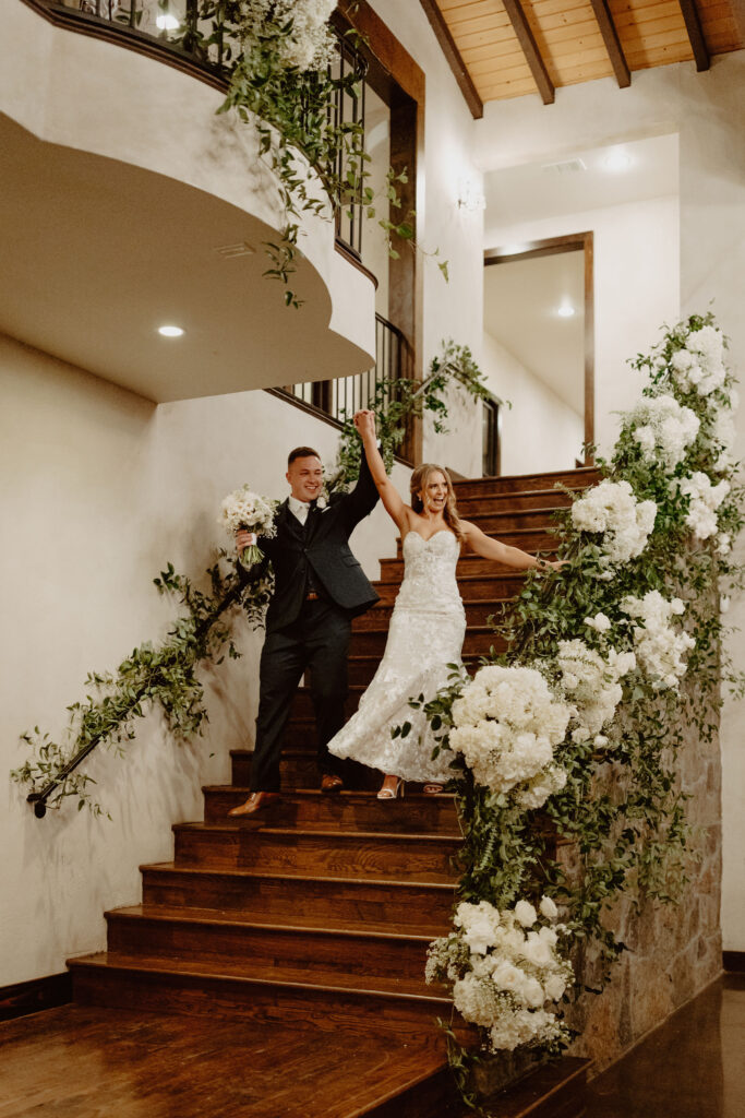 Bride and groom make a grand entrance down a floral-adorned staircase at Ma Maison, a premier wedding venue that's giving an idea of an average cost of wedding in Austin